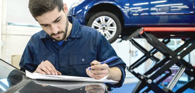 A updates a service log book in with a car behind him raised on a hydraulic lift