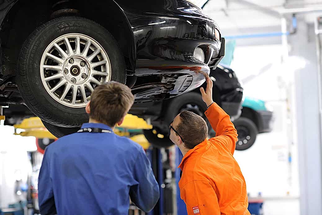 Two men under a car raised on a hydraulic lift peer speculatively