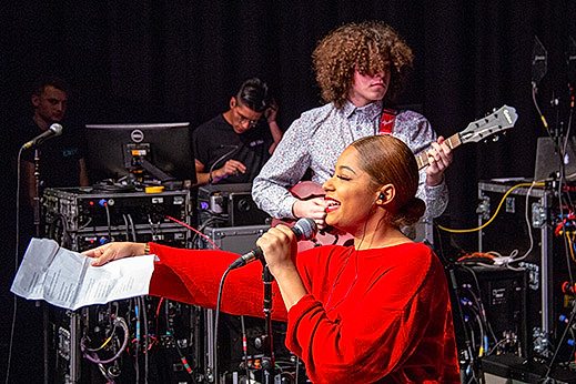 A young woman on stage smiles and gestures from the microphone stand to someone off camera