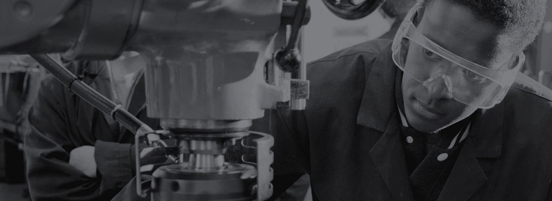 A young man in safety goggles peers closely into a piece of heavy machinery