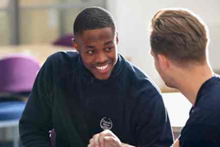 A young man turns smiling broadly at his friend in a sunny classroom