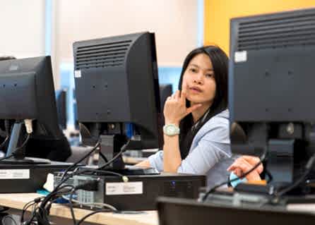 A young woman at a row of computers leans across to consult with her classsmate