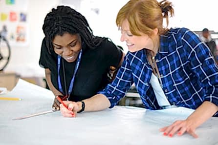 A student and her tutor discuss designs spread across a table in a brightly lit studio discussing